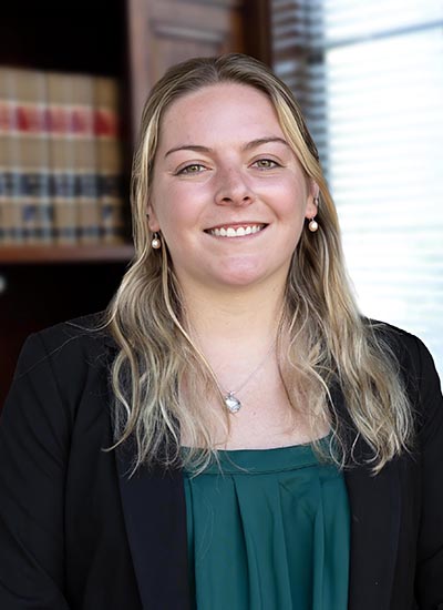 Smiling woman with shoulder-length blonde hair, wearing a green blouse and black blazer, in an office with a bookshelf in the background.